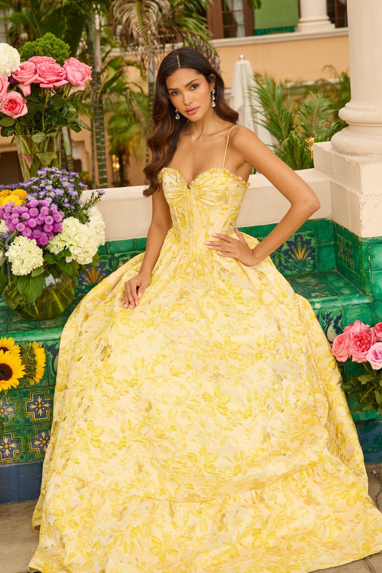 A woman in the Ava Presley 42312 long prom dress, featuring a yellow floral print and corset bodice, sits outdoors on a tiled ledge surrounded by colorful flowers and lush greenery.